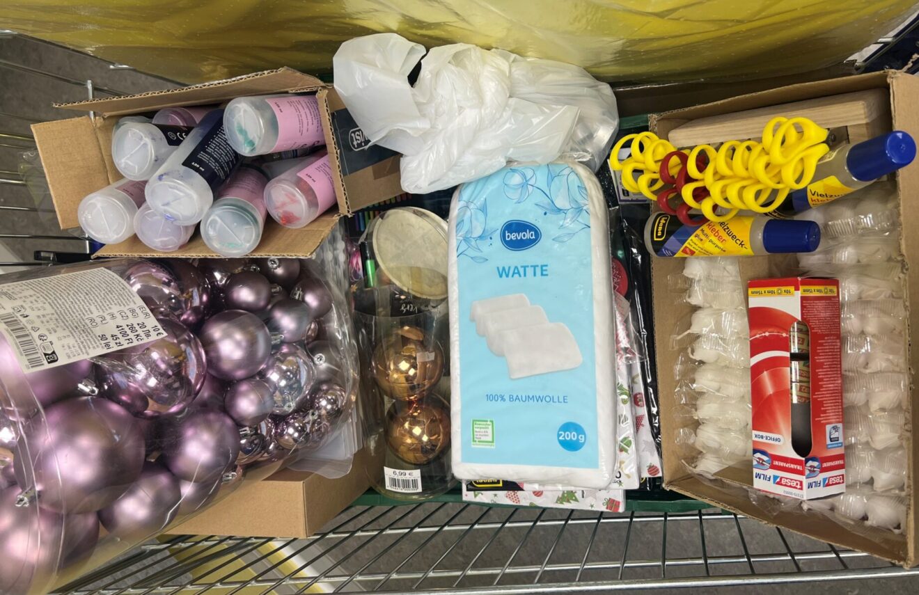 Ein Einkaufswagen, gefüllt mit rosafarbenem und goldenem Weihnachtsschmuck, Wattepads, gelben Bändern, Klebetuben, Plastikbechern mit Farbe und einer Schachtel mit Teelichtern.