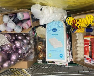 Ein Einkaufswagen, gefüllt mit rosafarbenem und goldenem Weihnachtsschmuck, Wattepads, gelben Bändern, Klebetuben, Plastikbechern mit Farbe und einer Schachtel mit Teelichtern.