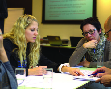 Personengruppe sitzt an einem Tisch in einem Besprechungsraum. Auf dem Tisch liegen Papierunterlagen, Stifte und Gläser mit Wasser. Im Hintergrund sind ein Laptop, ein Flipchart und eine Projektionsfläche mit Text erkennbar. Die Szene zeigt eine Arbeits- oder Diskussionsrunde.